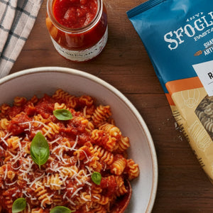 Prepared Sfoglini Classic Italian Pantry Set featuring dish with Garlic Radiators, tomato sauce, and basil leaves, accompanied by a jar of sauce and a package of Sfoglini pasta on a wooden table.